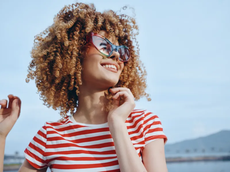 Portrait of a stylish young woman with voluminous curly hair, wearing a red and white striped tee and oversized sunglasses, smiling under a bright blue sky.