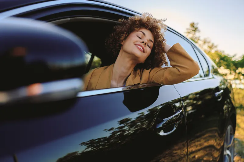 Woman enjoys freedom leaning from car window, eyes closed, smiling in a sunny outdoor scene with retro film color, relaxed mood, casual travel vibe, candid urban countryside.