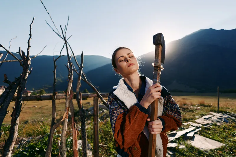 Woman outdoors countryside mountains holding a wooden tool, wearing sweater and shearling coat with sunlight on face, calm rural scene showing peaceful seasonal lifestyle and relaxation.