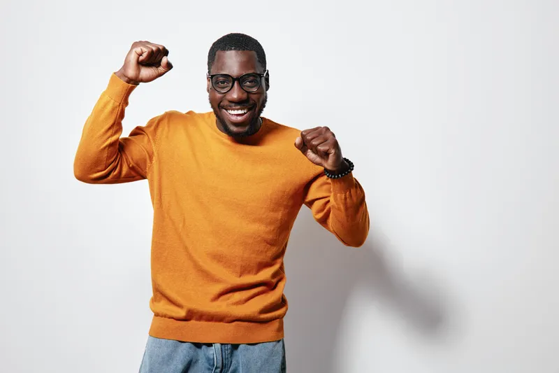 African American man with dark skin wearing glasses and orange sweater celebrating success with raised fists and smiling on white background. Casual style and positive emotion.