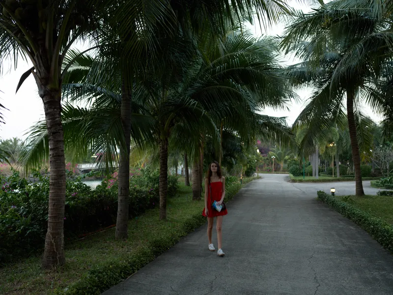 Young woman in red dress walking on tropical palm tree pathway during daytime with relaxed mood and casual style.