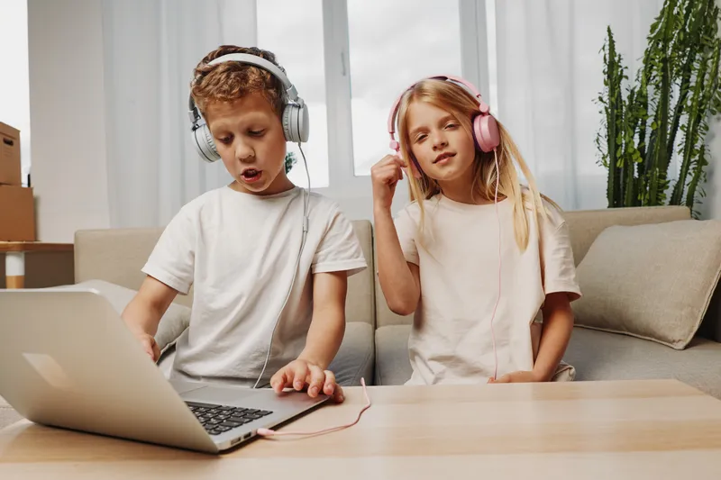 Two children wearing headphones enjoy music on a laptop in a cozy living room. The boy sings passionately while the girl listens with a relaxed expression. Concept for childhood joy and entertainment.