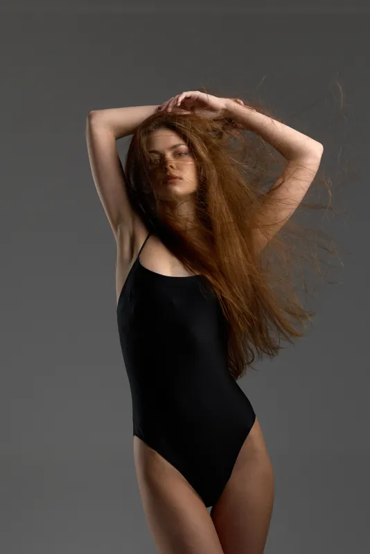 Woman with long red hair posing in black bodysuit against gray background with flowing hair and soft lighting