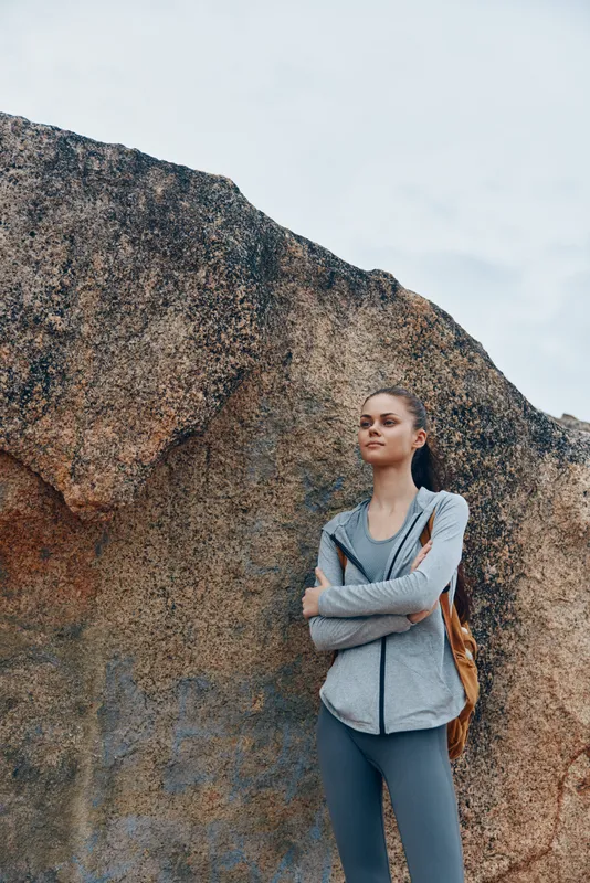 Young woman in athletic wear with a stylish backpack, confidently standing against a rocky background, promoting an active lifestyle and outdoor fitness