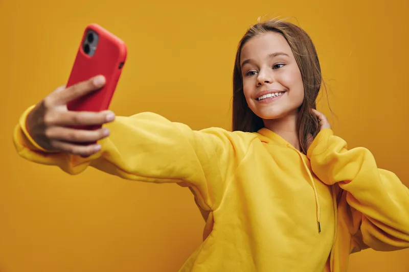 Girl in yellow hoodie taking selfie with red smartphone, smiling and posing on yellow background. Young female enjoying social media moment with casual style and happy expression.