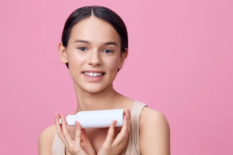 Young woman with a joyful expression holding a skincare product, wearing a simple top against a pink background, representing beauty and self care concepts