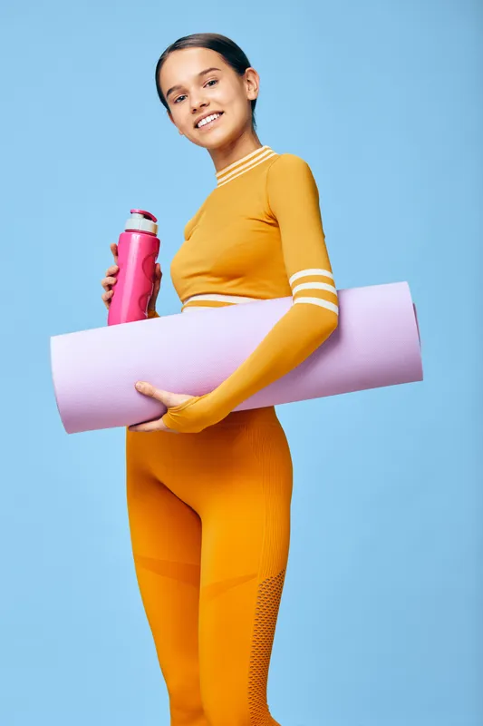 Young woman fitness athlete in bright orange activewear holding a pink water bottle and a rolled lavender yoga mat, smiling confidently in a minimalist light blue studio background.