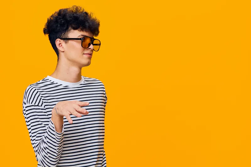 Stylish young man with curly hair wearing sunglasses and a striped shirt poses confidently against a vibrant orange background, showcasing casual fashion and cool attitude.