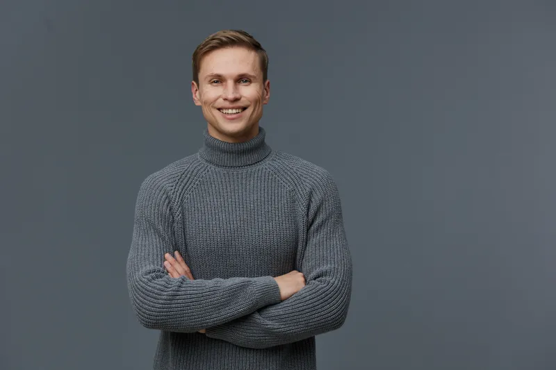 Smiling young man wearing gray turtleneck sweater posing confidently on plain gray background studio portrait. Casual fashion, friendly expression, cheerful mood, everyday style, male model, relaxed s