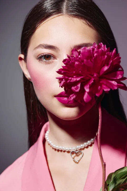 Young woman with glowing skin and bright lipstick holds dark pink flower covering half of her face. Creative beauty portrait with fashionable makeup and elegant pearl necklace.