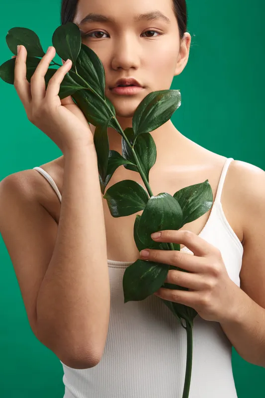 Asian woman holds a leafy stem near her face in a studio with a vivid green backdrop, conveying calm focus and contemporary beauty through natural pose and soft lighting.