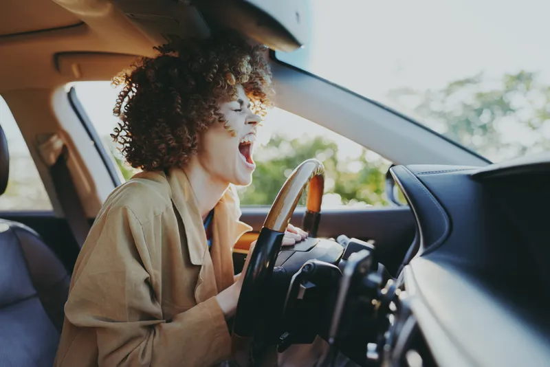 A young woman with curly hair shouts joyfully from the driver's seat while the car interior glows with retro film color, capturing a candid moment of carefree travel