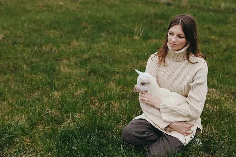 Calm woman in a cream sweater sits on grass holding a white goat kid. Natural daylight and open field create a gentle lifestyle scene of care, companionship, and rural harmony.
