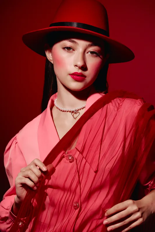 Young woman with rosy makeup wearing a bright hat and layered pink fashion clothes poses against red background. Concept of bold beauty, creative fashion and confidence.