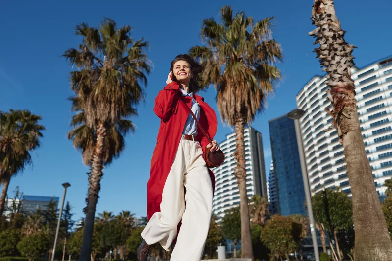 A stylish young woman strides through a sunny urban park lined with tall palm trees, wearing a red coat over light clothes and feeling confident and joyful.