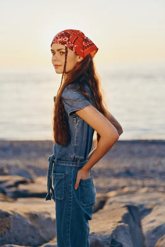 lifestyle woman in modern boho-western denim street style wearing red bandana with film color effect by the seaside during golden hour