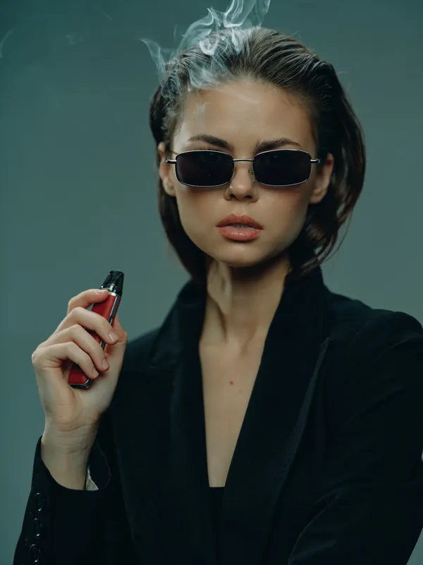 Stylish woman with sunglasses vaping electronic cigarette with smoke, wearing black outfit in blue background