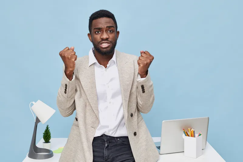 Confident young African American man in business casual attire celebrating success with clenched fists, standing in modern office space with laptop, desk organizer, and plant on pastel blue background