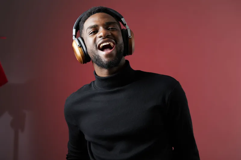 Smiling young Black man enjoying music with headphones, wearing a black turtleneck against a red background, conveying happiness and relaxation