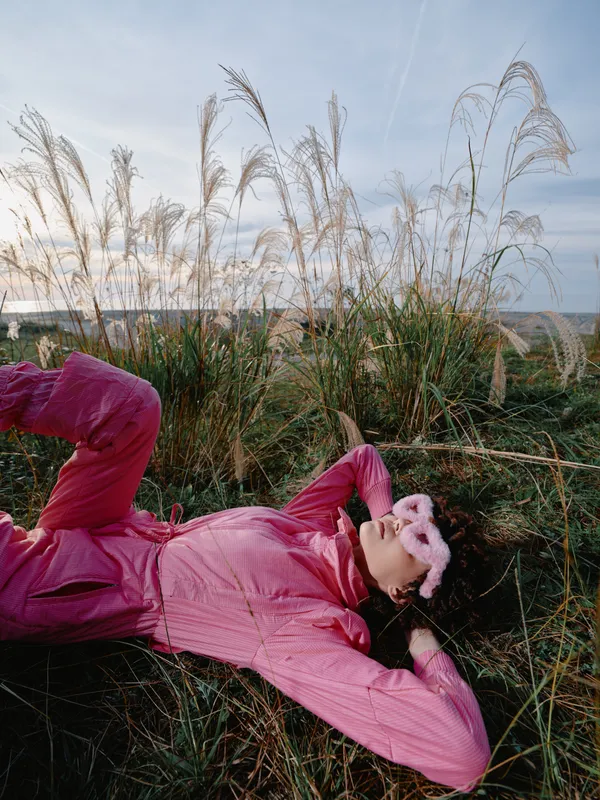 Woman lying in tall grass at dusk in a pink jacket, relaxing in a wide natural field with breeze and soft light, calm mood, outdoor leisure and rustic scenery.