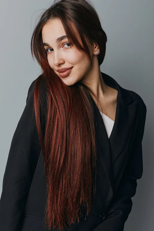 Young woman with long brown hair wearing black blazer posing and smiling on gray background. Portrait of confident female model with natural makeup and earrings in studio light for fashion.