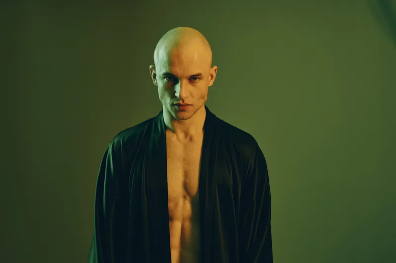 A contemplative man in a black robe stands against a softly lit background, embodying a sense of introspection and calmness. The image features light tones and shadow play.