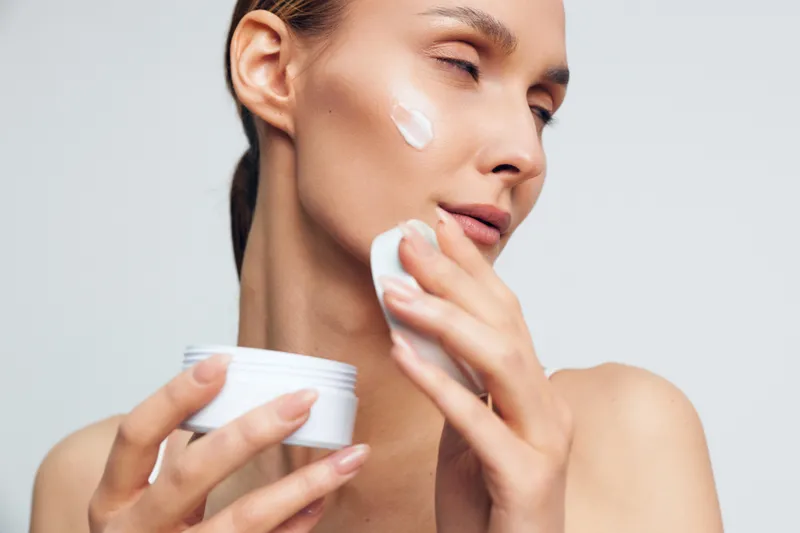 Woman skincare, face cream, applying moisturizer with closed eyes, holding jar and cotton pads, smooth skin, calm beauty routine, studio neutral background, daily care