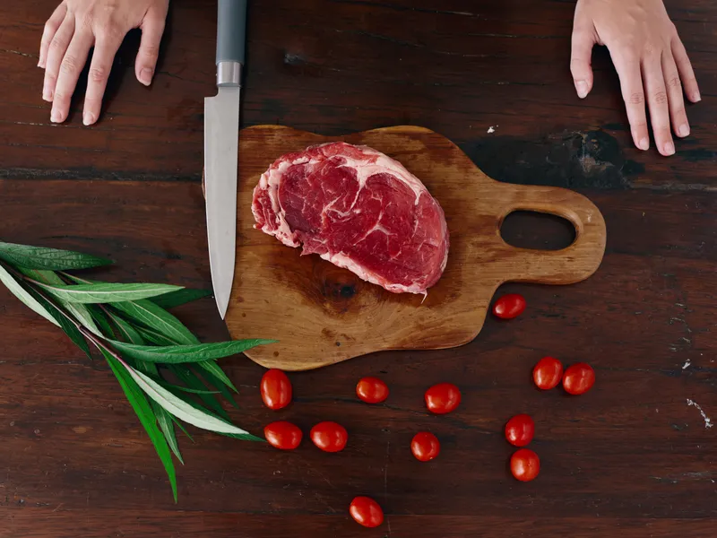 Женщина с ножом в руке нарезает свежее мясо для стейка на жарку на кухне