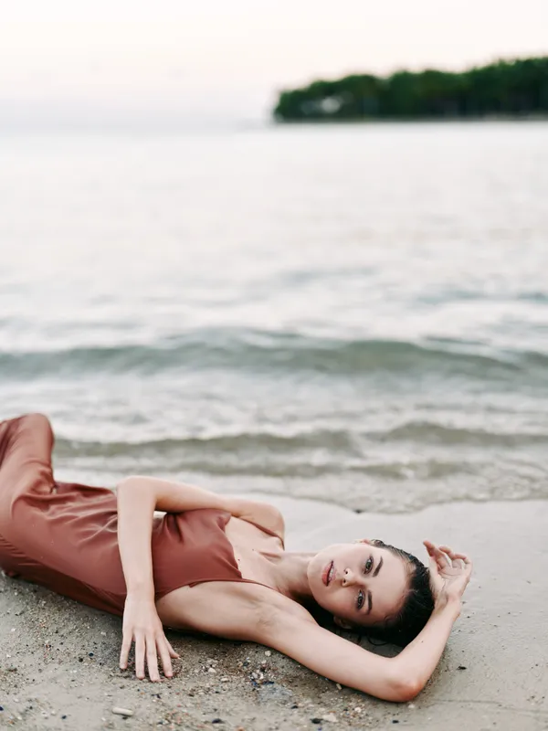 A graceful woman lies on a sandy beach in a rust brown dress, stretched along the shore as gentle waves lap nearby, creating a calm, intimate moment.