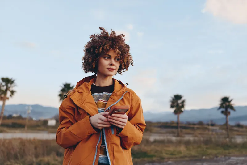 Woman lifestyle outdoors, curly hair, orange jacket, holding coffee cup, near palm trees and mountains under soft sunset light, relaxed casual travel mood, fresh air confidence