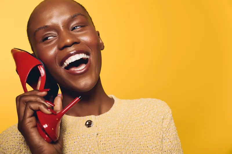 Happy African American woman with dark skin laughs holding red high heel shoe near face. Yellow background, bright clothing, joyful mood. Concept for fashion, beauty, happiness, and style.