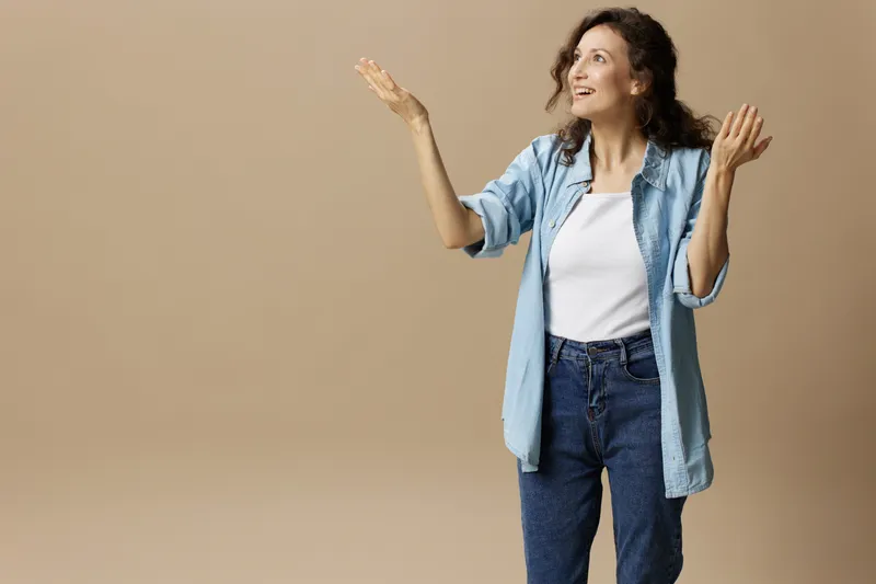 Веселая счастливая кудрявая красивая девушка в джинсах и повседневной рубашке указывает обеими руками вверх на свободное место для рекламы, позируя изолированно на бежевом пастельном фоне