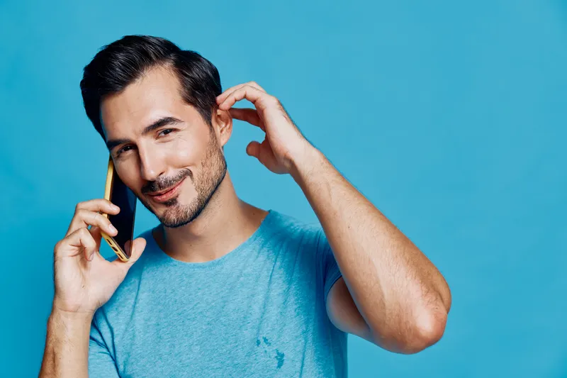 Full body smiling happy young man wearing blue t-shirt looking at camera while talking on mobile phone against solid blue background. Communication lifestyle concept.