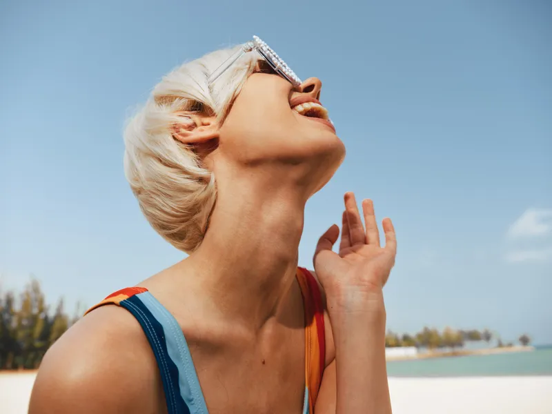 Sunlit moment of a carefree woman on a sunny beach, tilting her head back, swimsuit straps visible, sunglasses perched on her forehead beneath a clear blue sky.
