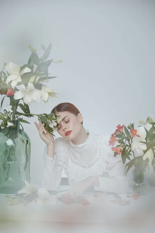 Serene young woman in white blouse surrounded by fresh flowers, evoking tranquility and beauty in a soft, pastel colored setting