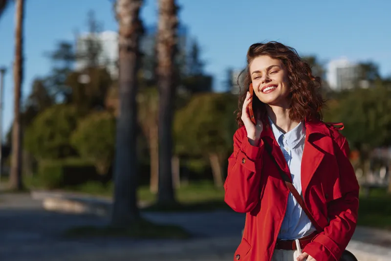 Smiling woman outdoors in a bright red coat, enjoying warm sunlight, relaxed posture, soft breeze, lush greenery, blue sky, candid portrait with natural colors.