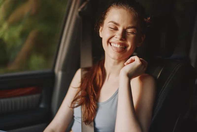 Happy young woman with long brown hair smiling in a car, enjoying a carefree moment during a summer road trip adventure in a vibrant green setting