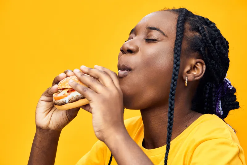 Cheerful girl enjoying a tasty burger against a vibrant yellow background, celebrating delicious food with joy Perfect for food and lifestyle themes