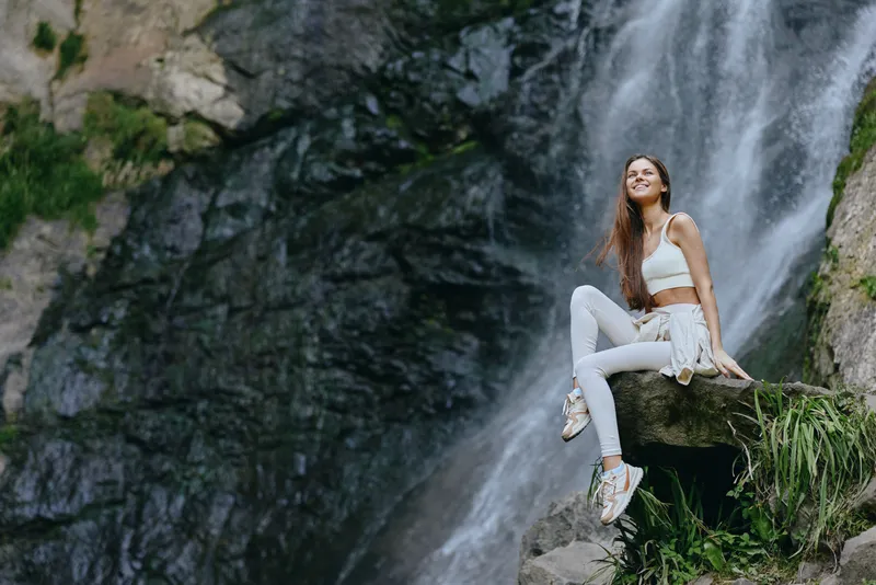 young woman sitting near waterfall, wearing white sportswear, smiling happily outdoors in nature, surrounded by rocks and flowing water.