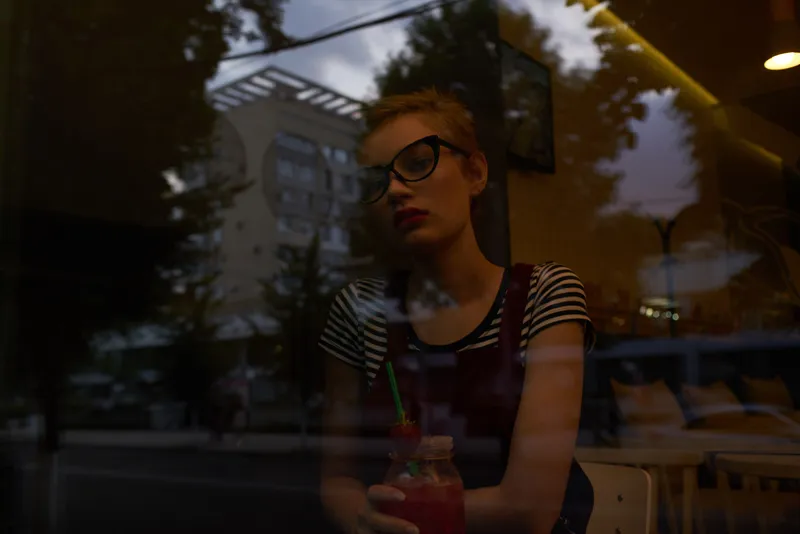 Женщина смотрит в окно уличного кафе, потягивая коктейль, что передаёт атмосферу отдыха и образа жизни