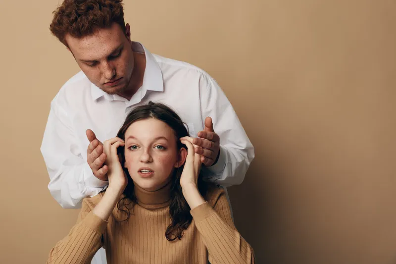 Young man in white shirt massages temples of woman in brown sweater. Both have serious expressions, isolated on beige background. Concept of stress relief, relaxation, and wellness.