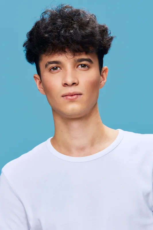 young man with curly hair wearing white t-shirt looking confident against light blue background, youthful male portrait with natural skin tone and casual style