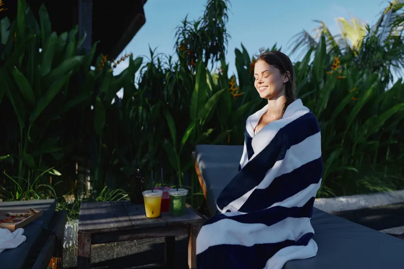 A young woman wrapped in a striped blanket sits outdoors smiling with closed eyes near tropical plants on a sunny day. Lifestyle concept of relaxation and comfort.