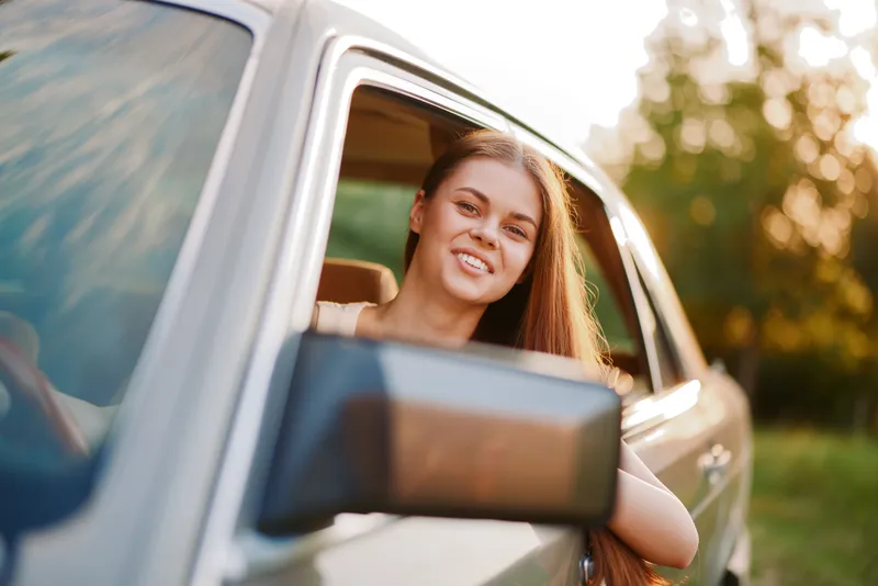 Young woman enjoying a joyful moment leaning out of a car window during sunset, showcasing her long hair and sunny smile amidst a green background.