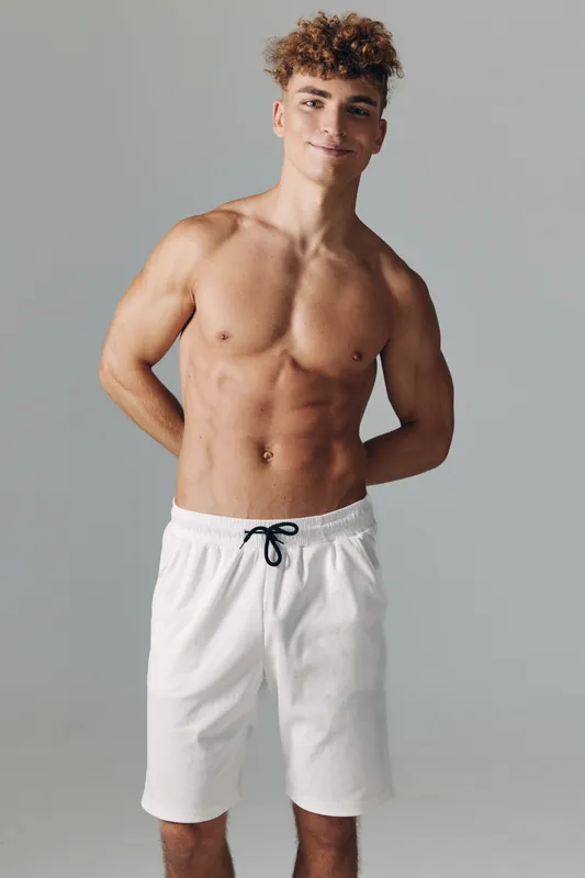 Young fit man standing shirtless and smiling in white shorts with black drawstring against grey background isolated studio portrait of confident sporty model with curly hair and muscular body