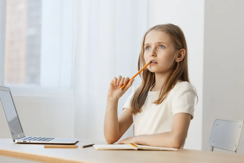 Pensive young girl with long hair, pondering while studying at a desk with a laptop and notebook, capturing a moment of deep thought and curiosity