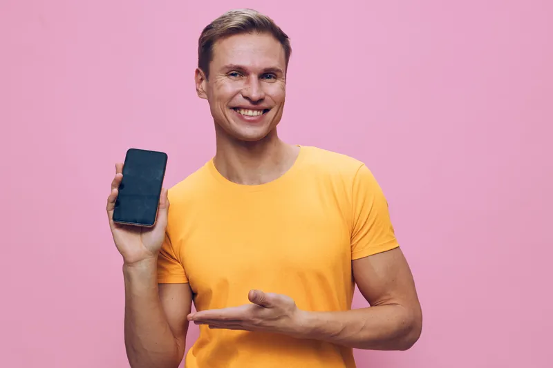 Smiling young caucasian man holding smartphone in yellow t-shirt on pink background digital nomad concept technology connection communication