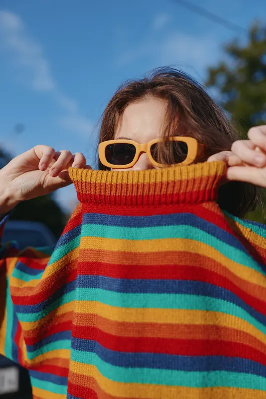 woman striped sweater sunglasses playful outdoor portrait of a young adult in her mid twenties, brunette with yellow shades pulling colorful knit over her face, casual street fashion, sunny vibe