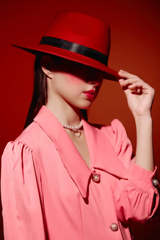 Fashionable woman in pink jacket and red hat tilts hat visor with hand, lips painted red. Creative beauty image with dramatic studio light and contrast. Fashion and style concept.