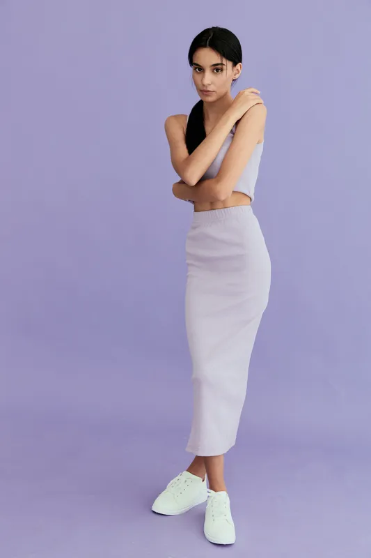 Young woman standing against a lavender background wearing a light gray matching top and skirt with white sneakers, posing with crossed arms and a calm expression.